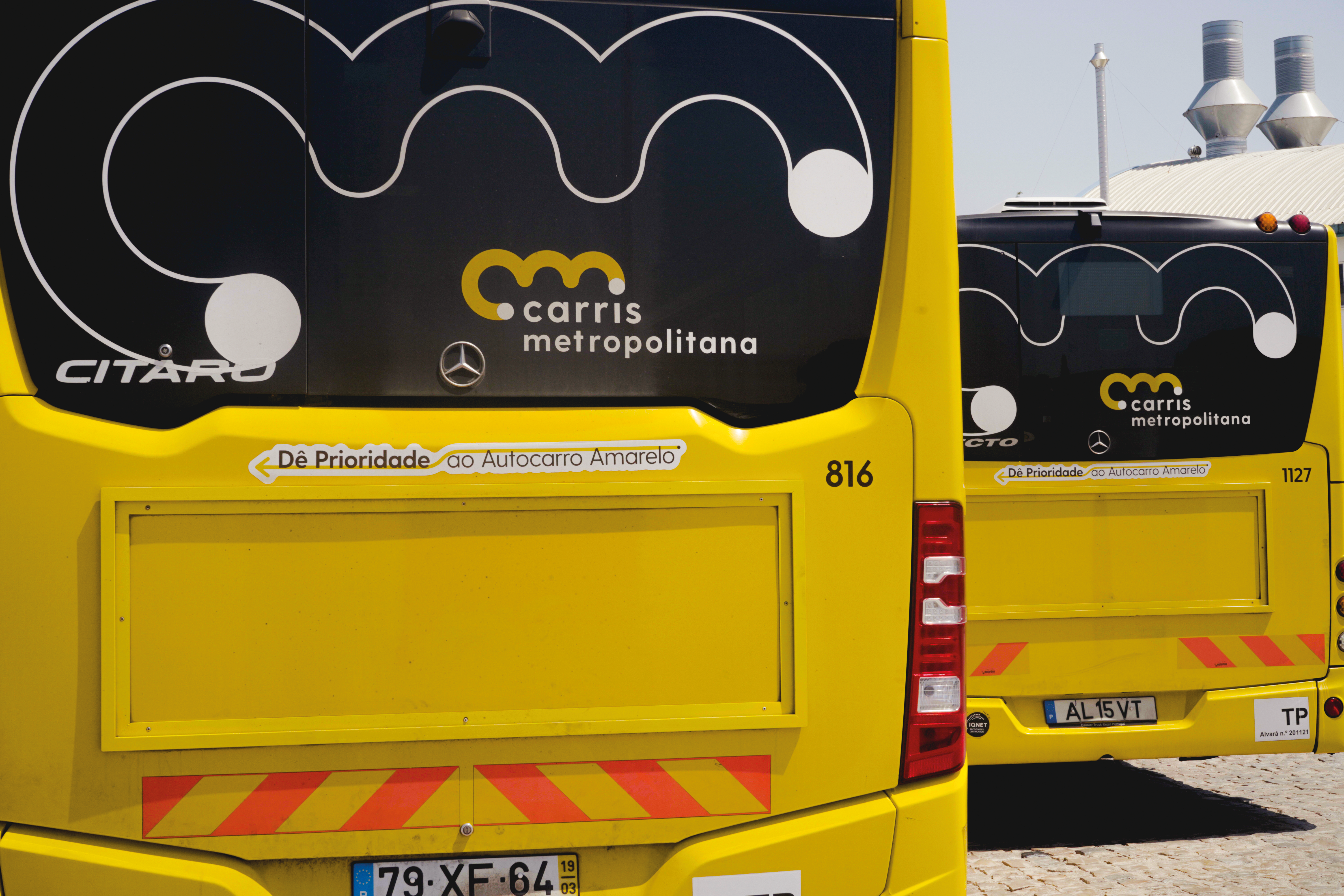 Parte traseira de dois autocarros amarelos da Carris Metropolitana estacionados, com o logótipo visível e a frase “Dê prioridade ao autocarro amarelo”